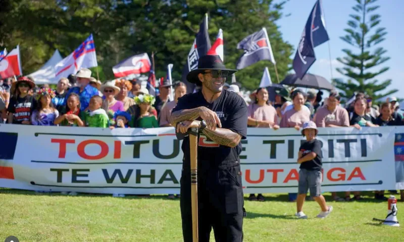 A man in black stands in front of a large crowd holding a banner that reads "TOITŪ TE TIRITI ME TE WHAKAPUTANGA." People in the crowd wave flags and wear hats and sunglasses on a sunny day with trees in the background.