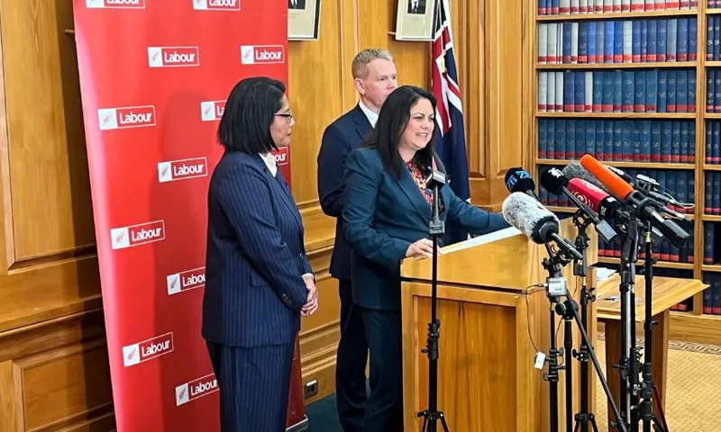 Ayesha Verrall stands at a podium, speaking into microphones in front of a Labour Party banner. Beside her are Barbara Edmonds and Chris Hipkins.