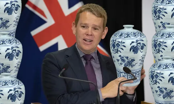 A man in a suit speaks at a podium, holding a blue and white porcelain vase. Several similar vases surround him. A partially visible flag with blue, red, and white colors is in the background.