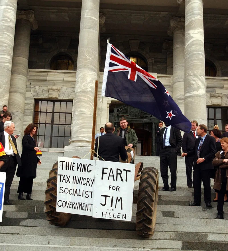 A man on a tractor with protest signs and a New Zealand flag is parked on parliament steps, surrounded by people in formal clothing. The signs read "THIEVING TAX HUNGRY SOCIALIST GOVERNMENT" and "FART FOR JIM AND HELEN.