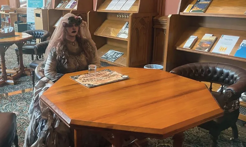 A woman wearing tattered Victorian garb sits at a table in parliament's library, with an ouija board in front of her.
