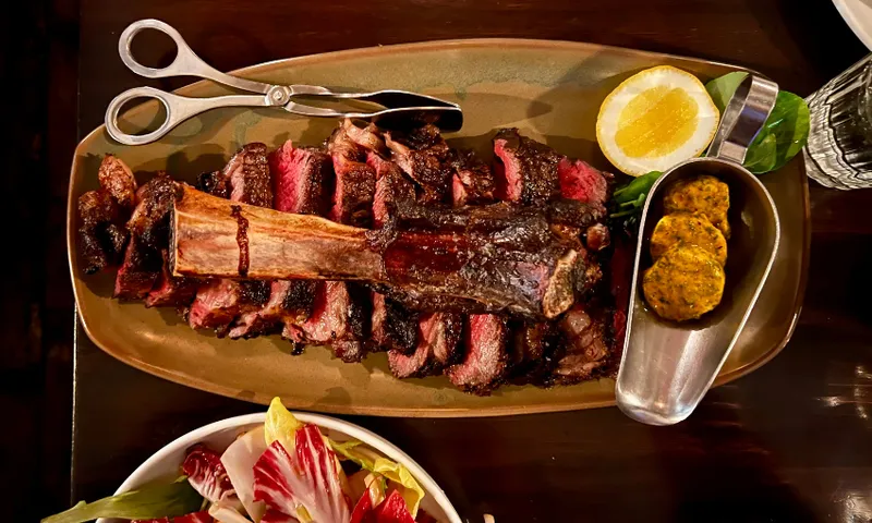 Sliced grilled steak served on a platter with a large bone, accompanied by a halved lemon, two slices of compound butter, scissors, and a glass of water. A salad with leafy greens is partially visible.