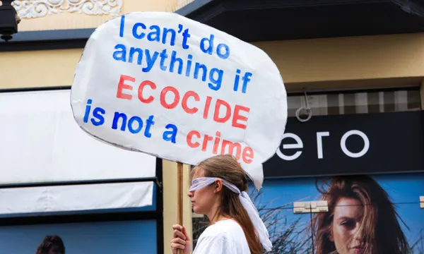 A woman wearing a white t-shirt and white blindfold holds a placard reading "I can't do anything if ecocide is not a crime"