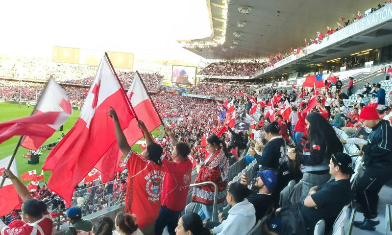 Fans in a stadium wave large red and white flags, filling the stands with color and energy. Many spectators wear red shirts, showing support, while the seats and field are packed with enthusiastic crowds.