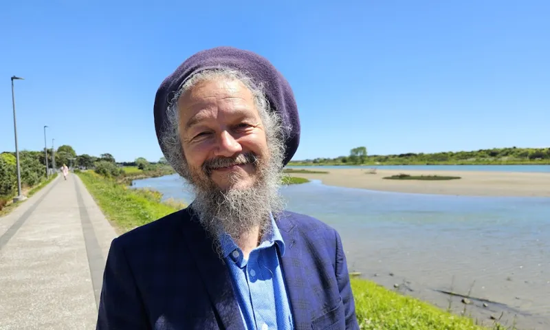 A smiling man with a grey beard and a purple hat stands by a river on a sunny day, with a walkway and greenery in the background under a clear blue sky.