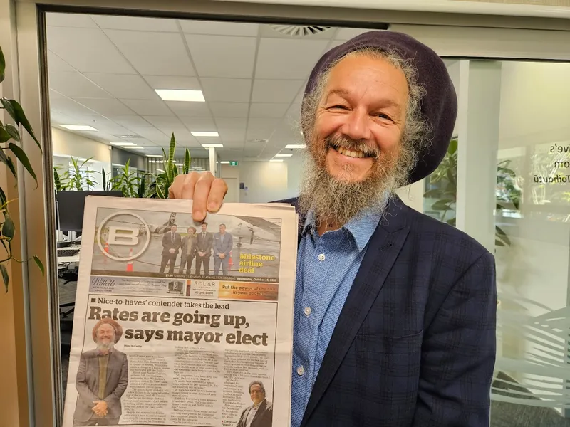 A smiling man with a gray beard and dreadlocks, wearing a dark blazer and hat, holds up a newspaper featuring his photo on the front page with the headline "Rates are going up, says mayor elect.