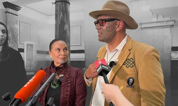Two Māori individuals with traditional facial tattoos are being interviewed by reporters holding microphones. The man wears a tan jacket, hat, and sunglasses; the woman wears a maroon blouse. The background is black and white.