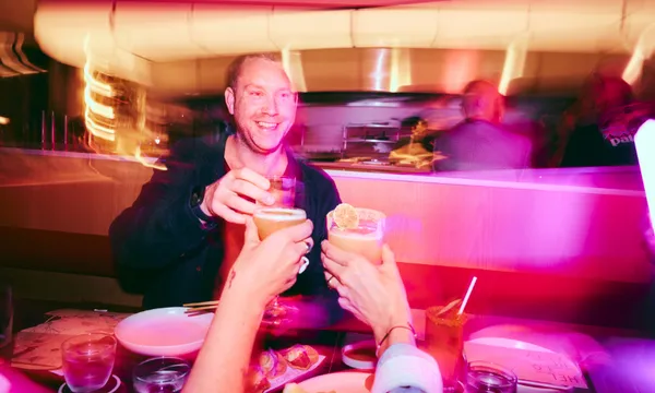 A group of people raise cocktails and glasses in a celebratory toast at a restaurant table, surrounded by bright, colorful lighting and a blurred background.