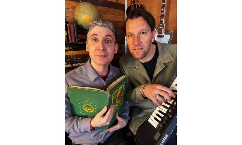 A photo of two men, one holding an open book and the other playing on a keyboard.
