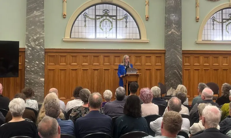 Helen Joyce, wearing a blue power suit, speaks to a crowd of people who have the back of their heads facing the camera.