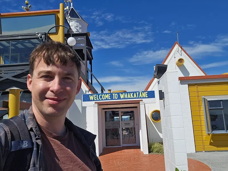 A man with brown hair smiles at the camera in front of a building with a sign that reads “Welcome to Whakatāne” against a blue sky.