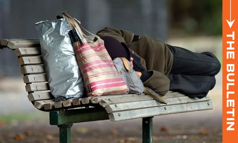 A person lies on a park bench, resting their head on a backpack, with bags beside them. The image is overlaid with an orange vertical banner reading "THE BULLETIN" on the right side.