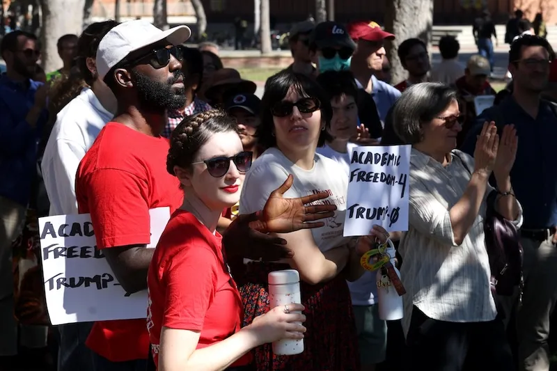 A diverse group of people at an outdoor rally, some holding signs that read "ACADEMIC FREEDOM = TRUMP." Most are wearing sunglasses, and they appear attentive and engaged in the event.