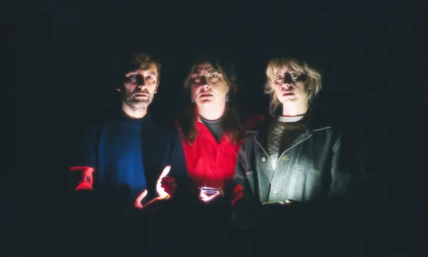 Three people stand closely together in the dark, illuminated dramatically from below by a soft light. Their faces are serious, and the background is completely black, creating a mysterious and intense mood.