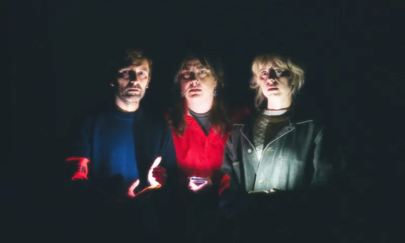Three people stand closely together in the dark, illuminated dramatically from below by a soft light. Their faces are serious, and the background is completely black, creating a mysterious and intense mood.