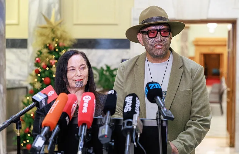 A woman and a man with tā moko stand at a podium with multiple microphones in front of them, speaking to the media indoors. A decorated Christmas tree is visible in the background.