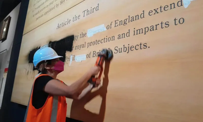 A person wearing a hard hat, pink mask, and orange safety vest uses a power tool to erase text from a large wall display titled "Article the Third.