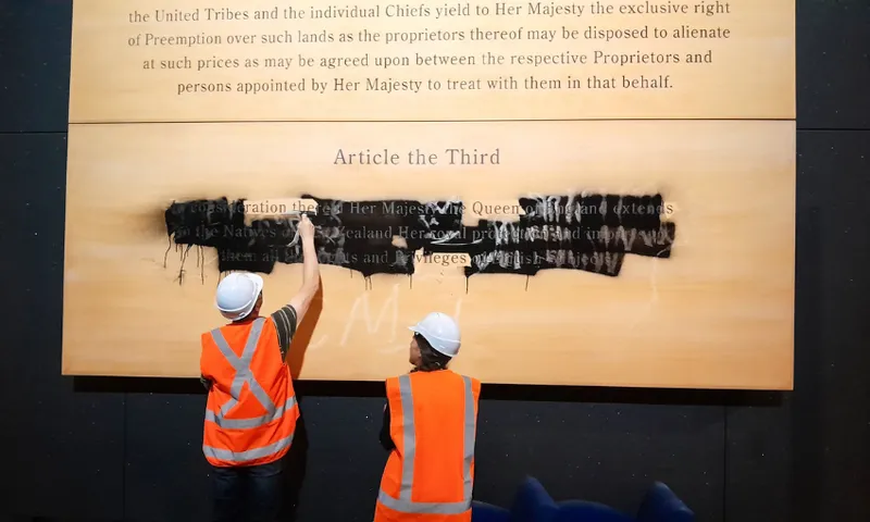 Two people in orange safety vests and white helmets paint over English text on a large display panel under the heading "Article the Third," partially obscuring the words with black paint.