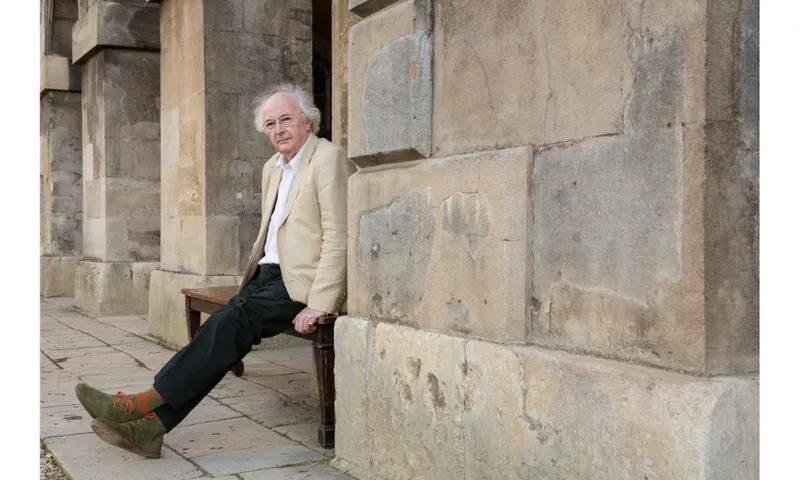 A photo of the writer Philip Pullman who is in a cream blazer and black pants and is leaning against a stone buildling.