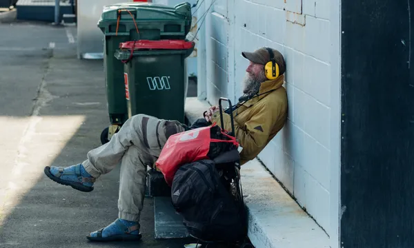 A man with a gray beard wearing headphones, a brown jacket, and sandals relaxes against a white wall outdoors, surrounded by bags and near green trash bins.