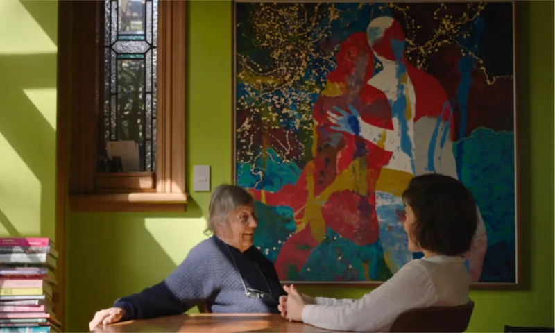 an older woman speaks to a younger woman at a table. a colourful large painting hangs on the green wall behind them