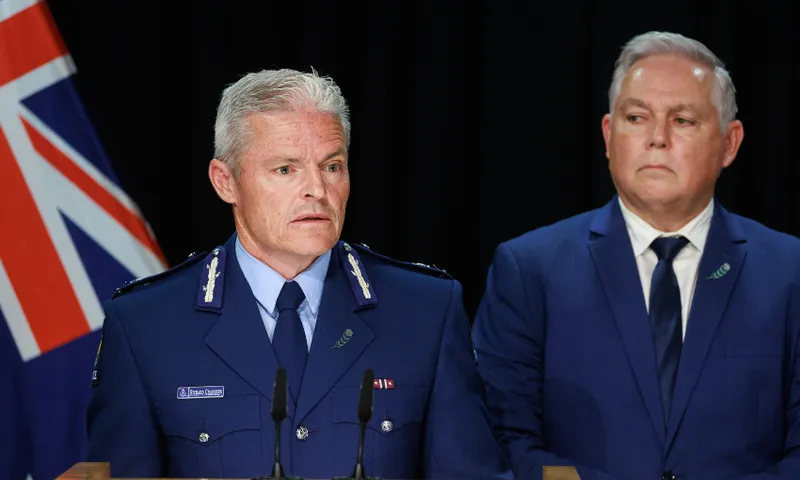 Two men in blue suits stand at a podium, one in a police uniform speaking, with a New Zealand flag visible in the background against a black curtain.