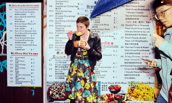 A woman in a colorful dress eats from a bowl with chopsticks in front of a large Chinese restaurant menu. A man with glasses drinks from a cup on the right side of the image. Various dishes are pictured on the menu.