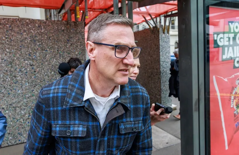 A man wearing glasses and a blue plaid jacket walks outdoors near a glass wall and a sign, with people and red structures in the background.