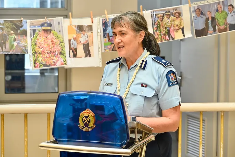 A policewoman in uniform speaks at a podium. Behind her, photos of police officers and events are displayed on a string. She wears a white lei and stands indoors, smiling as she addresses an audience.