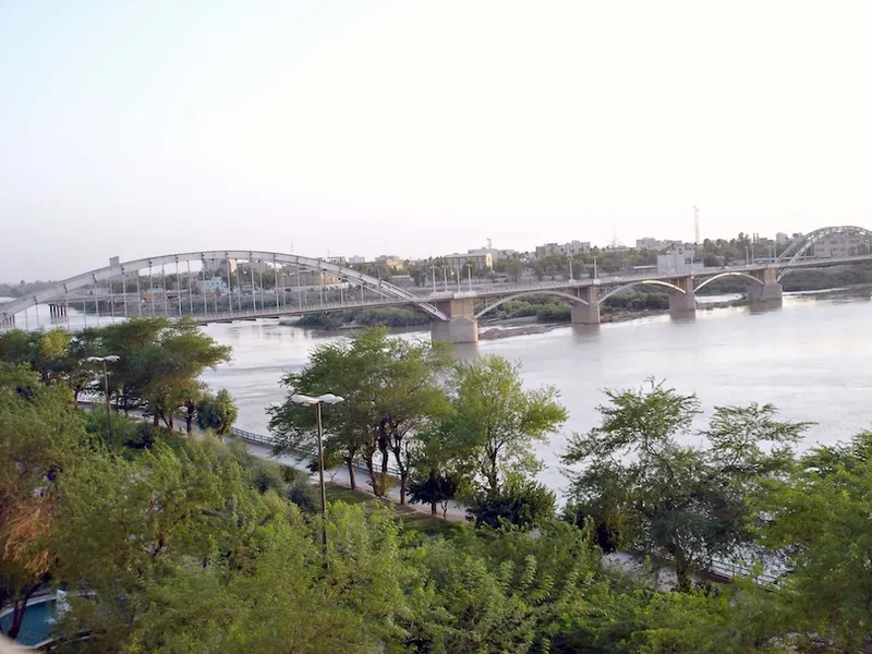A large arched bridge spans a wide river, surrounded by lush green trees along the riverbank, with buildings visible in the distance under a pale sky.