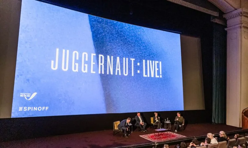 A wide shot taken inside of The Embassy theatre, with the big screen reading: "Juggernaut: Live!". Chris Bishop, Chris Wikaira, Linda Clark and Toby Manhire sit beneath the screen.
