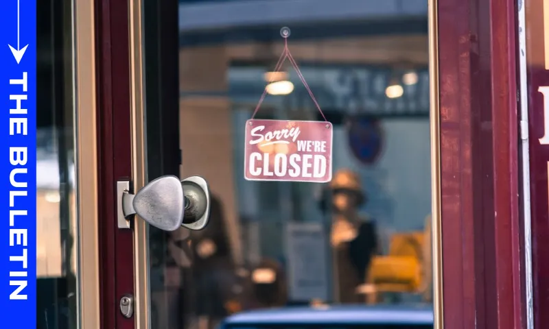 A glass door with a "Sorry, we're closed" sign hanging on it. Reflections and a blurred interior are visible through the glass. A blue vertical banner on the left reads "THE BULLETIN.
