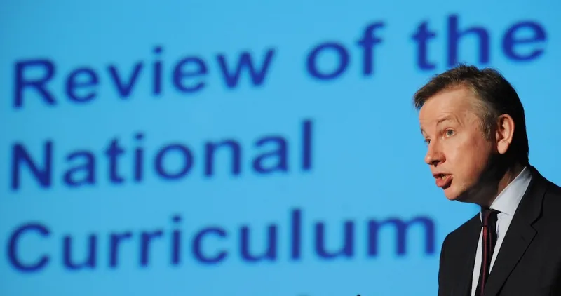 A man in a suit speaks in front of a blue screen displaying large text that reads “Review of the National Curriculum.”.