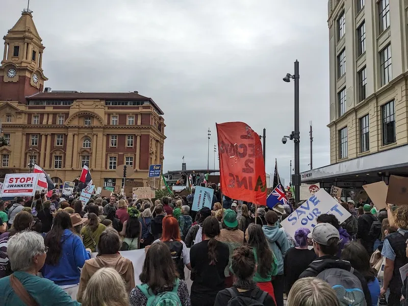 lots of people holding signs with auckland's ferry terminal visible in the background