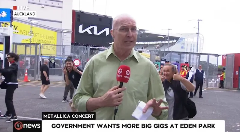 TVNZ reporter Simon Mercep talks to camera as Metallica fans in the background wave to the camera