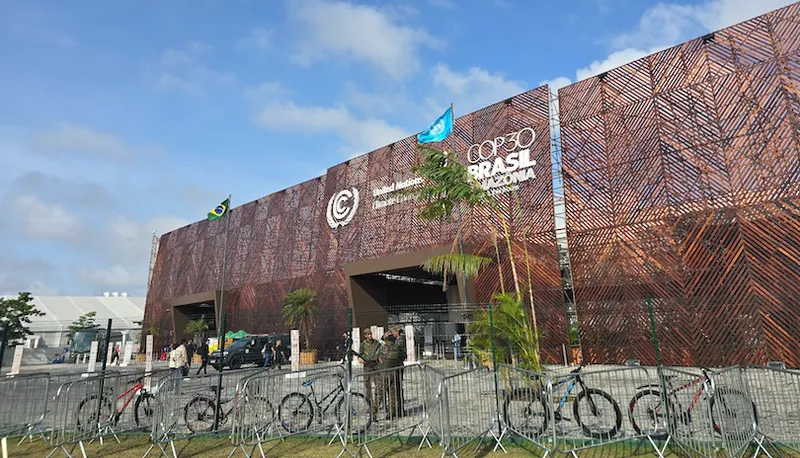 Large modern building with a geometric patterned facade, displaying "COP30 Brasil Amazônia" and climate conference logos. Bicycles are parked along fences in front, and flags, including Brazil’s, are visible on the roof.