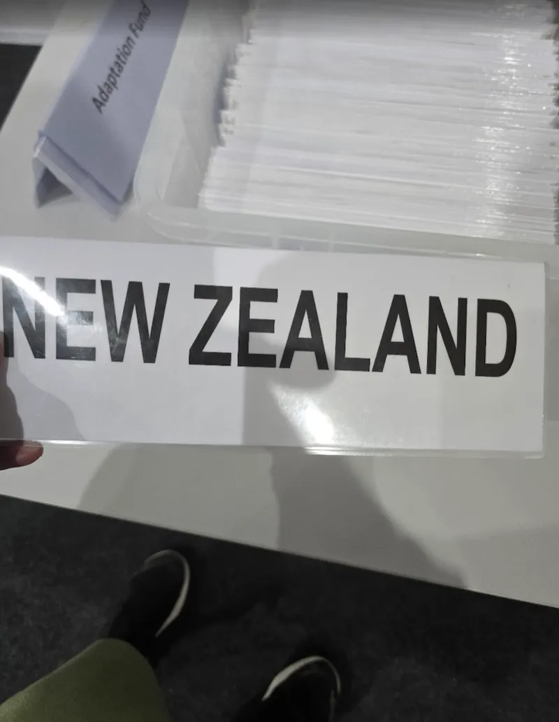A person is holding a sign that reads "NEW ZEALAND" in bold black letters. In the background, there are labeled folders, one of which reads "Adaptation Fund.