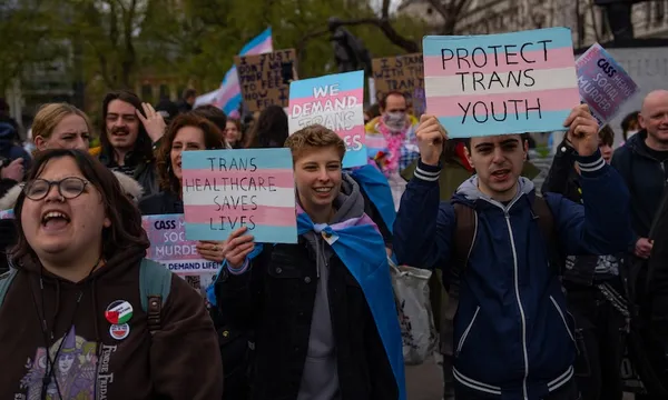 A group of people at a protest hold signs reading "TRANS HEALTHCARE SAVES LIVES" and "PROTECT TRANS YOUTH," with transgender pride flags visible. The mood is energetic and supportive.