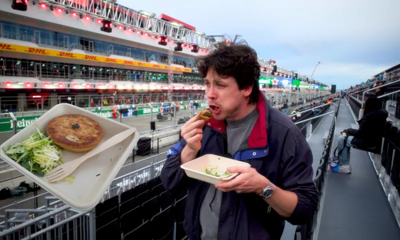 A man wearing a jacket stands in the stands of a grand prix track and is mid-bit of a pie
