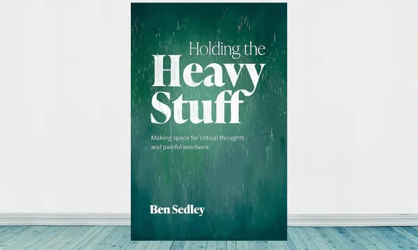 The cover of a book called Holding the Heavy Stuff: the cover is bottle green texture with white text. It sits on a background emulating the inside of a house with white walls and a wooden floor.