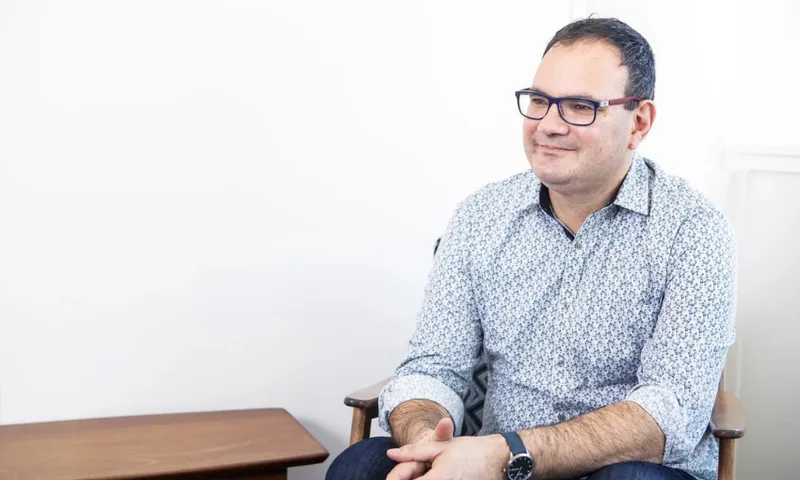 A photograph of a white man with glasses and a shirt on sitting at a table, on a chair. He is smiling.