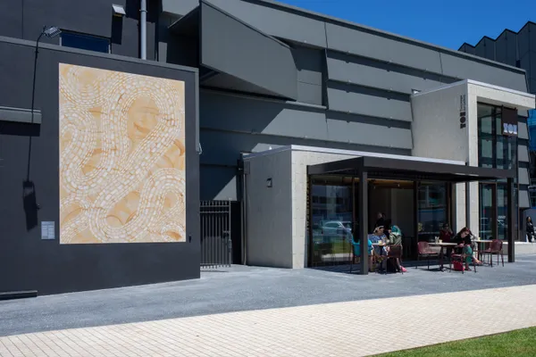 A large abstract mural is displayed on the exterior wall of a modern gray building. People sit at tables outside a café near the entrance, enjoying a sunny day.