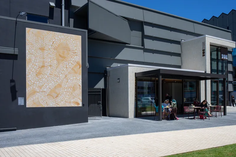 A large abstract mural is displayed on the exterior wall of a modern gray building. People sit at tables outside a café near the entrance, enjoying a sunny day.