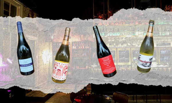 Four wine bottles with different labels are displayed in a row over a torn paper effect, with a dimly lit, stylish bar and shelves of glasses in the background.