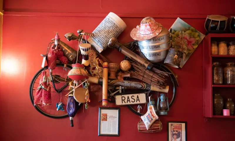 A red wall decorated with a vintage bicycle adorned with baskets, hats, musical instruments, a sign reading "RASA," framed photos, and jars filled with spices.