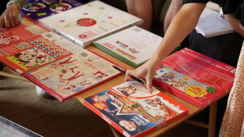 Several colorful advent calendars are spread out on wooden tables. One person is reaching out to touch or examine a box while others are seated nearby, some holding notebooks.