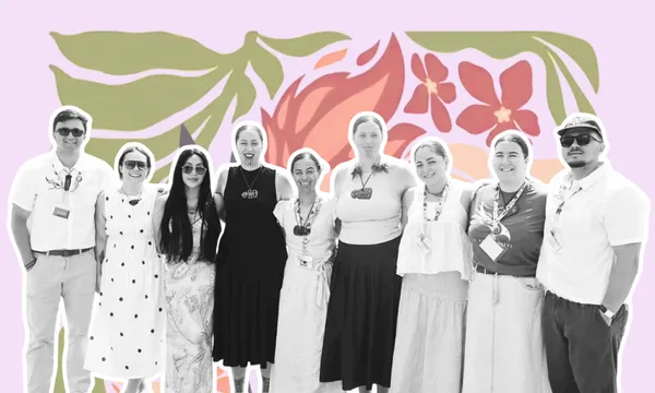 A group of nine people stand side by side, smiling, in front of a colorful floral-patterned background. The image is in black and white, except for the pastel and vibrant flowers behind them.