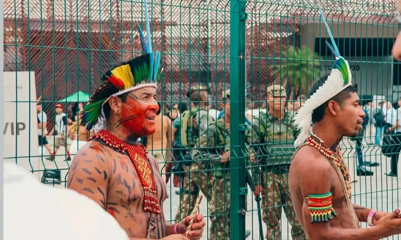 Two Indigenous men wearing traditional headdresses, face and body paint, and beaded jewelry stand near a metal fence. In the background, several uniformed soldiers are visible.