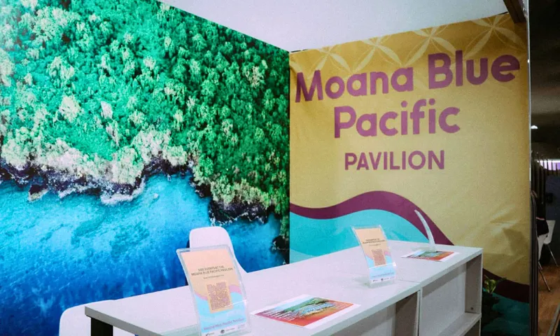 A booth with a sign reading "Moana Blue Pacific Pavilion" stands beside a wall displaying an aerial photo of lush green trees and blue ocean. Three clear display stands with brochures are on the white counter.