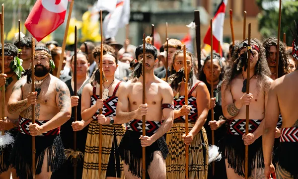 A group of Māori men and women dressed in traditional clothing stand together holding wooden spears during a cultural parade or ceremony, with flags and a crowd visible in the background.